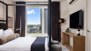 A hotel room with a bed, a desk with a TV, a bottle of wine, and a view of the city.