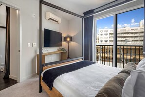 A modern hotel room with a large bed, a TV, a desk with a lamp, and a balcony with a city view. - Designer 2BR | Gym, Parking (White Plains)