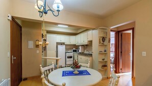 A dining area with a round table, chairs, and a chandelier. A kitchen is visible in the background with white cabinets and appliances.