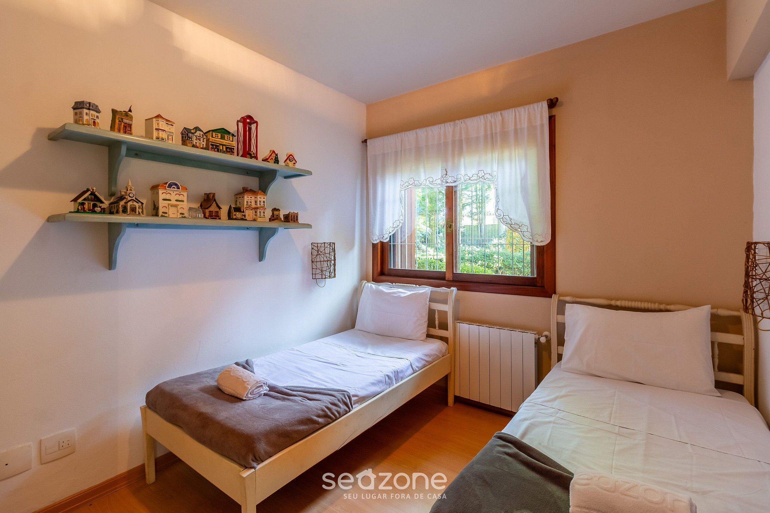 A small, cozy room with a single bed, a window with curtains, and shelves with model houses.