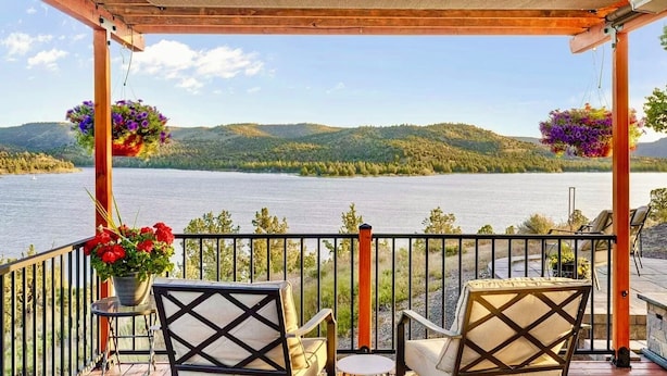 A covered outdoor seating area with two chairs and a table, overlooking a lake with hills in the background.