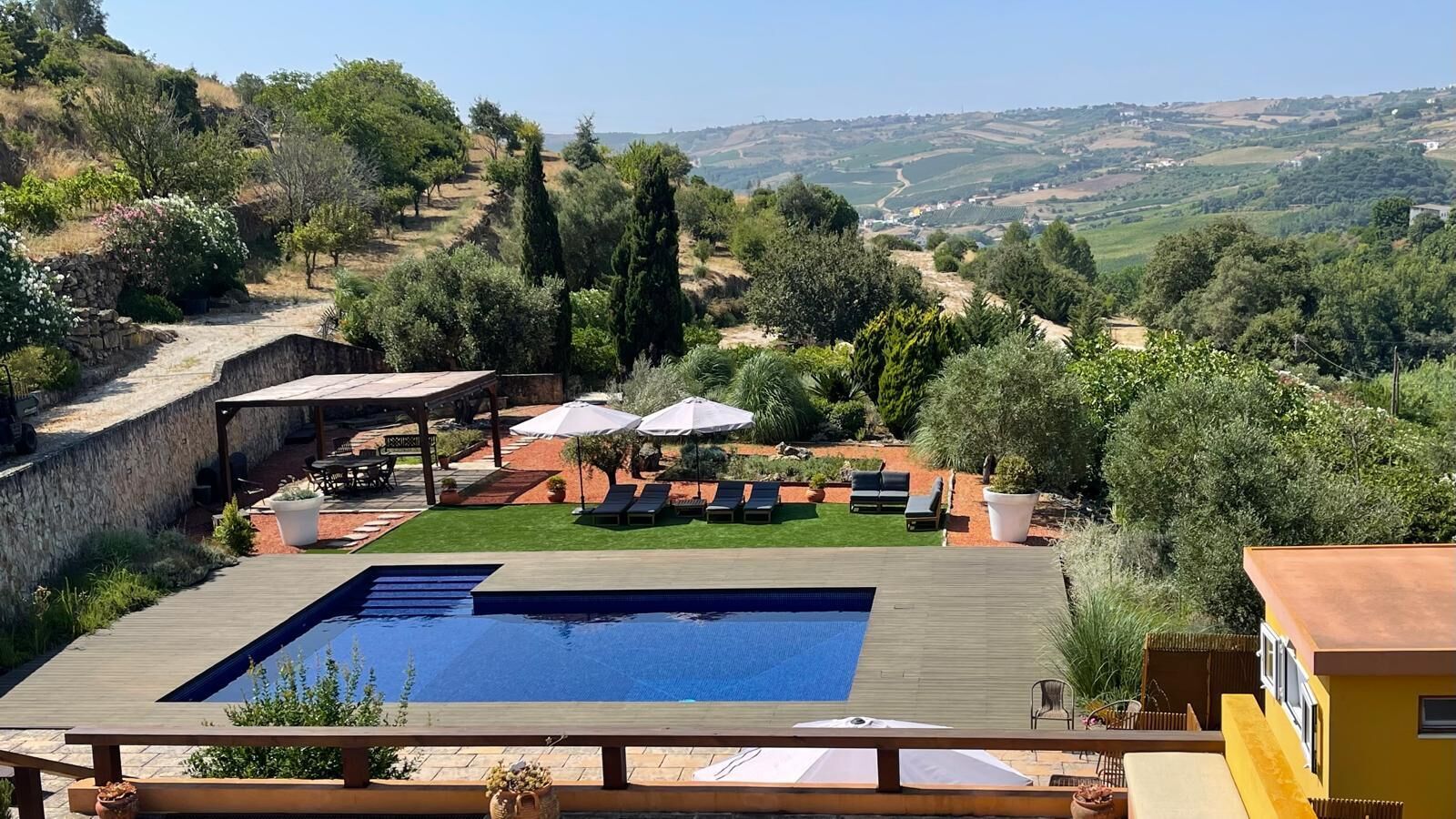 A swimming pool with a wooden deck, surrounded by lounge chairs and umbrellas, set against a hillside backdrop.