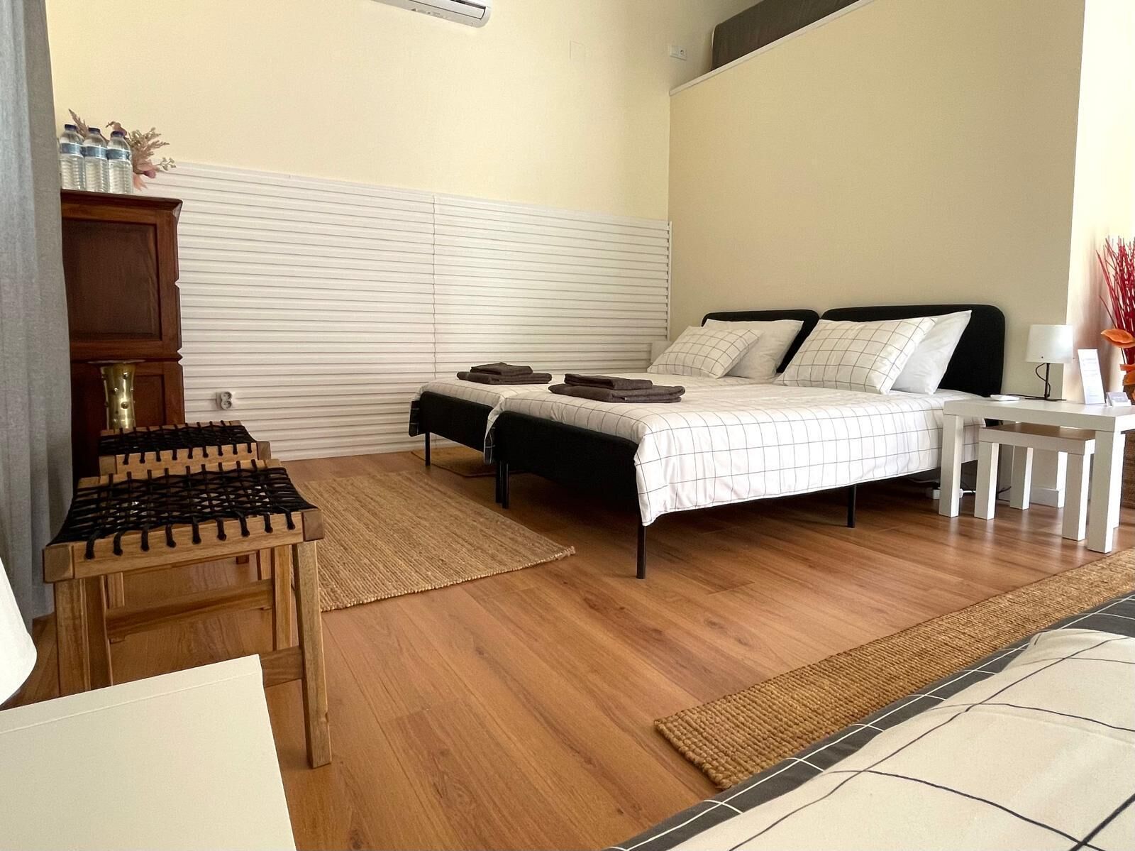 Un dormitorio con cama, mesitas de noche, una silla de madera, una pequeña mesa con estufa y una ventana con persianas.