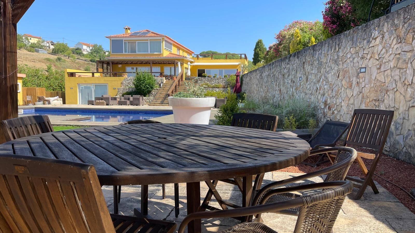 A patio with a round table and chairs, a stone wall, and a pool in the background.