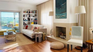 Een moderne woonkamer met een witte bank, een boekenkast, een open haard en een groot raam met uitzicht op de omgeving.