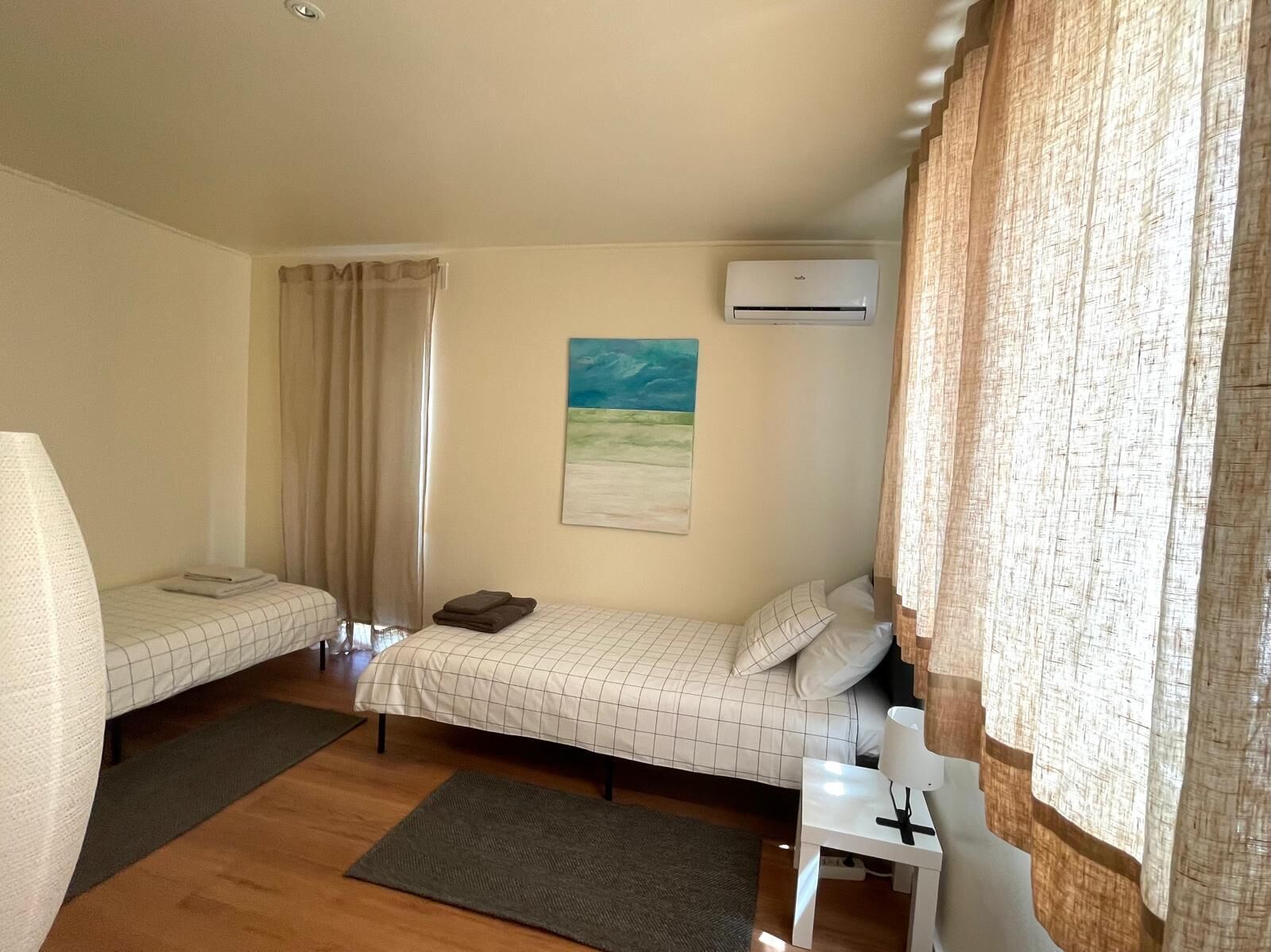 A small, well-lit room with two single beds, a small table, an air conditioning unit, and a painting on the wall.