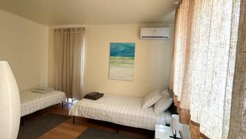 A small, well-lit room with two single beds, a small table, an air conditioning unit, and a painting on the wall.