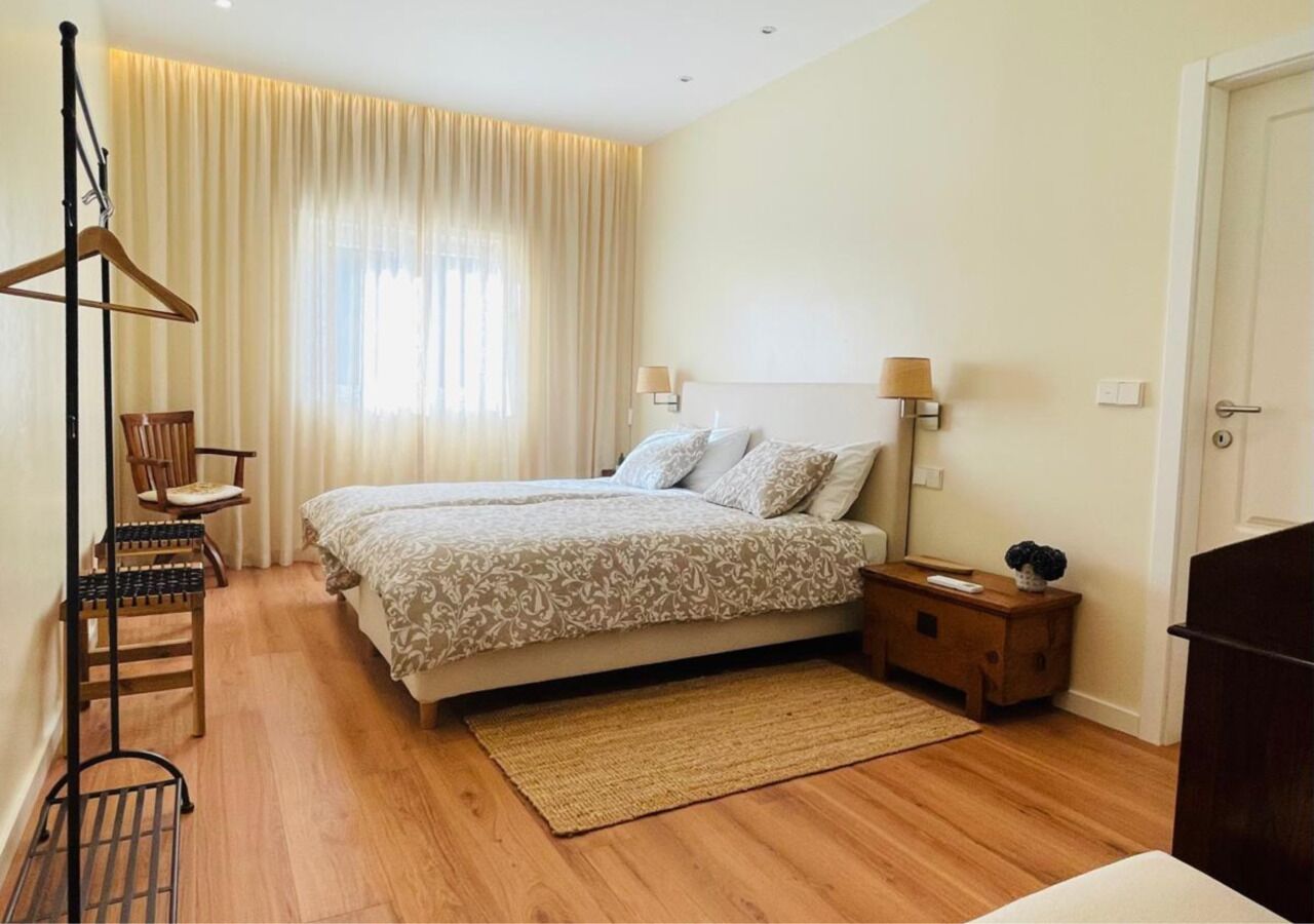 A bedroom with two beds, a wooden floor, a bedside table with a lamp, a chair, and a coat rack.