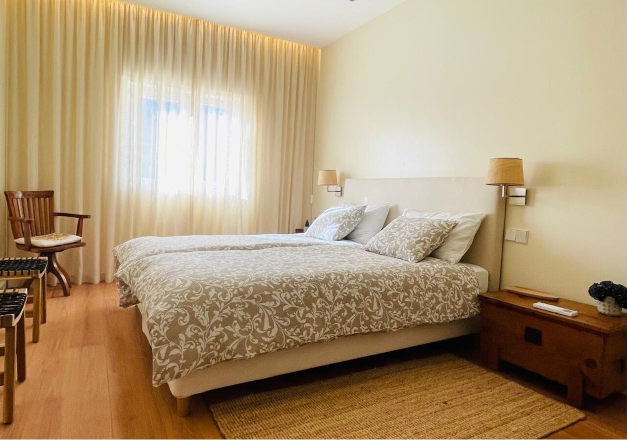 A double bed with patterned bedding, a wooden nightstand with a lamp, a chair, and a small table with a remote control.