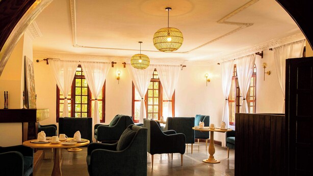 A room with a round table set for dining, several upholstered chairs, a small side table, and a large window with curtains. - The Hidden Valley Lodge (Arusha)