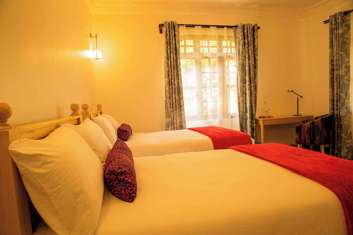 A hotel room with two beds, a desk with a lamp, and a window with curtains.