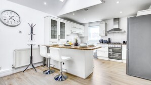 A modern kitchen with white cabinetry, a central island, bar stools, a clock, and a large window. - Wimbledon/Discount/Ensuite/Close to AELTC&Station (London)