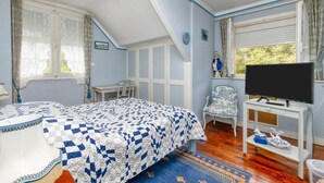 Un dormitorio con una cama, televisión sobre un mueble, una silla y una ventana con cortinas.