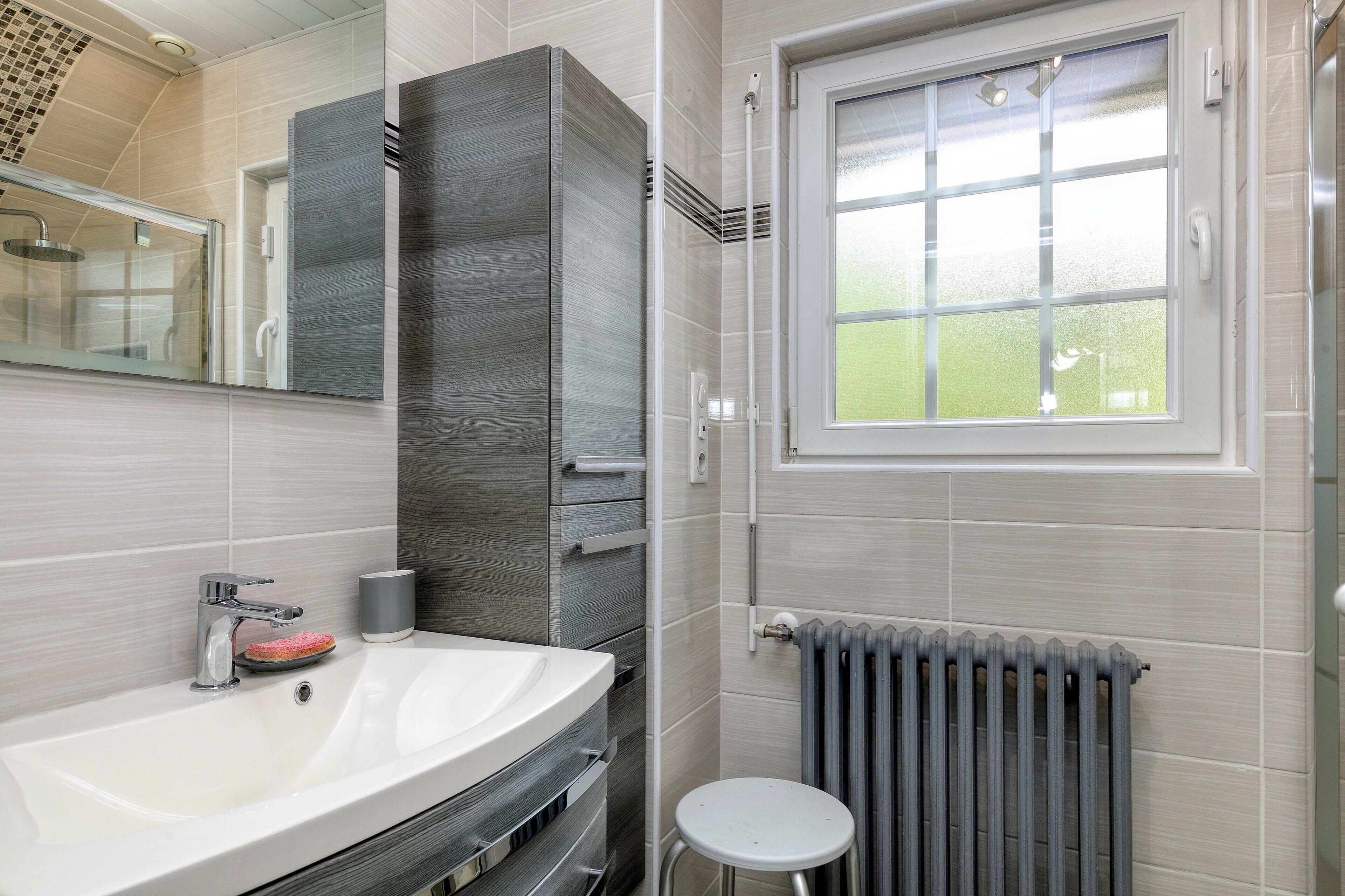 Une salle de bain moderne avec un lavabo blanc, une cabine de douche en verre, un radiateur et une fenêtre.
