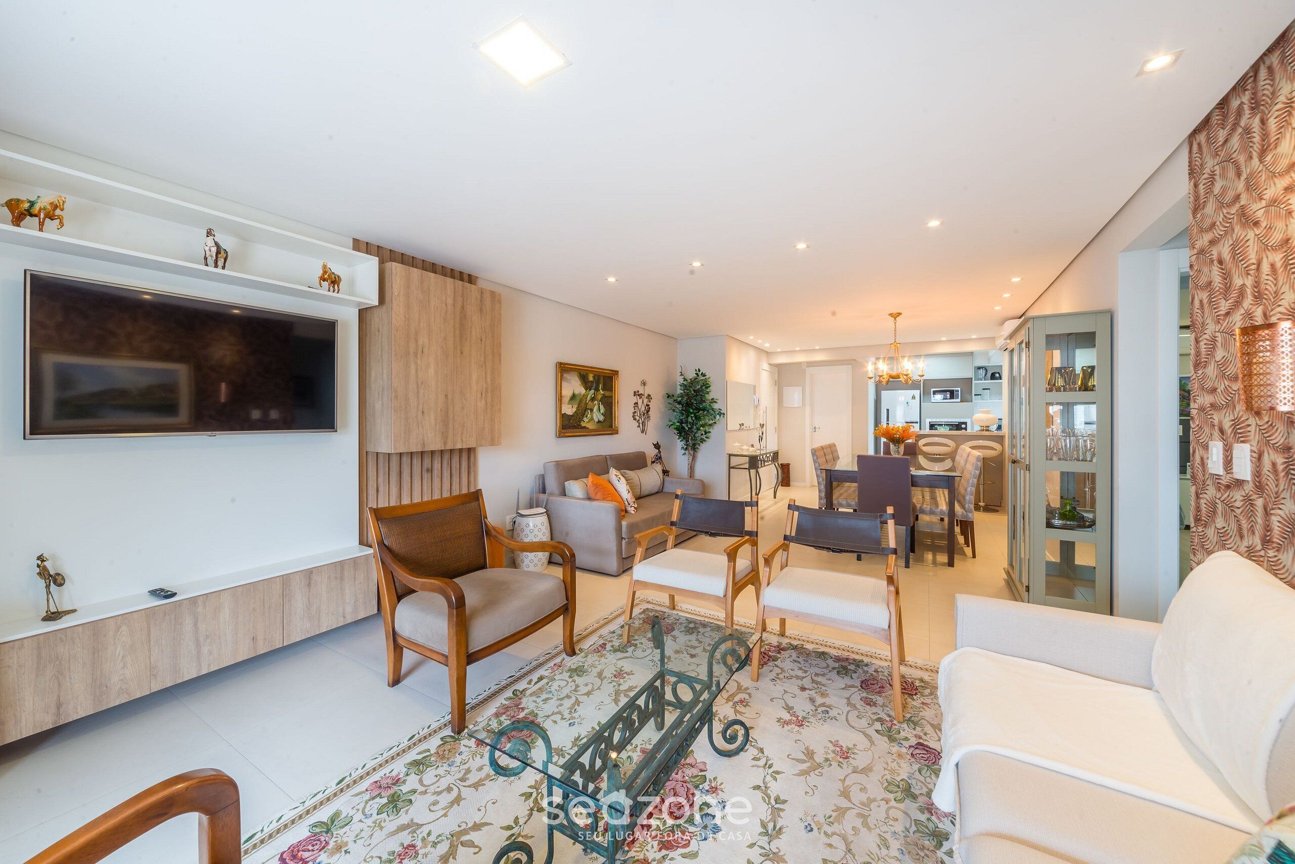 A modern living room with a flat-screen TV, wooden furniture, a dining area with chairs, and a patterned rug.