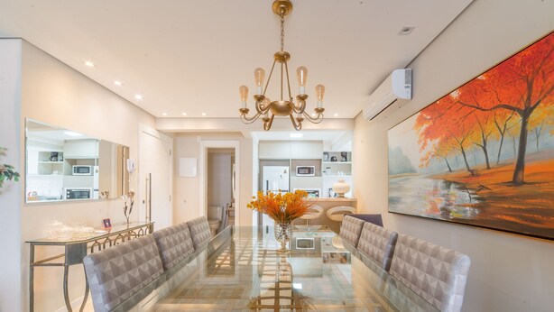 A modern dining area with a glass table, cushioned benches, a chandelier, and a painting of autumn trees. - NSA - Apartments Close to the Center of Canela RS (Canela)