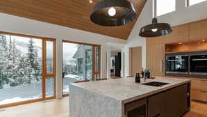 Une cuisine moderne dotée d’un îlot en marbre, de meubles en bois et de grandes fenêtres offrant une vue sur un paysage enneigé.