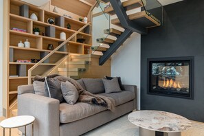 A modern living room with a sofa, a coffee table, a fireplace, a bookshelf, and a staircase. - Aurora Estate at Kadenwood (Whistler)
