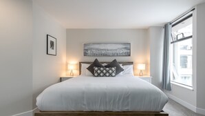 Une chambre à coucher moderne avec un lit surélevé en bois, deux lampes de chevet, un tableau encadré et une grande fenêtre avec des rideaux.