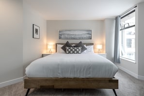 A modern bedroom with a wooden platform bed, two bedside lamps, a framed picture, and a large window with curtains. - Big Mauna at the Marquise (Whistler)