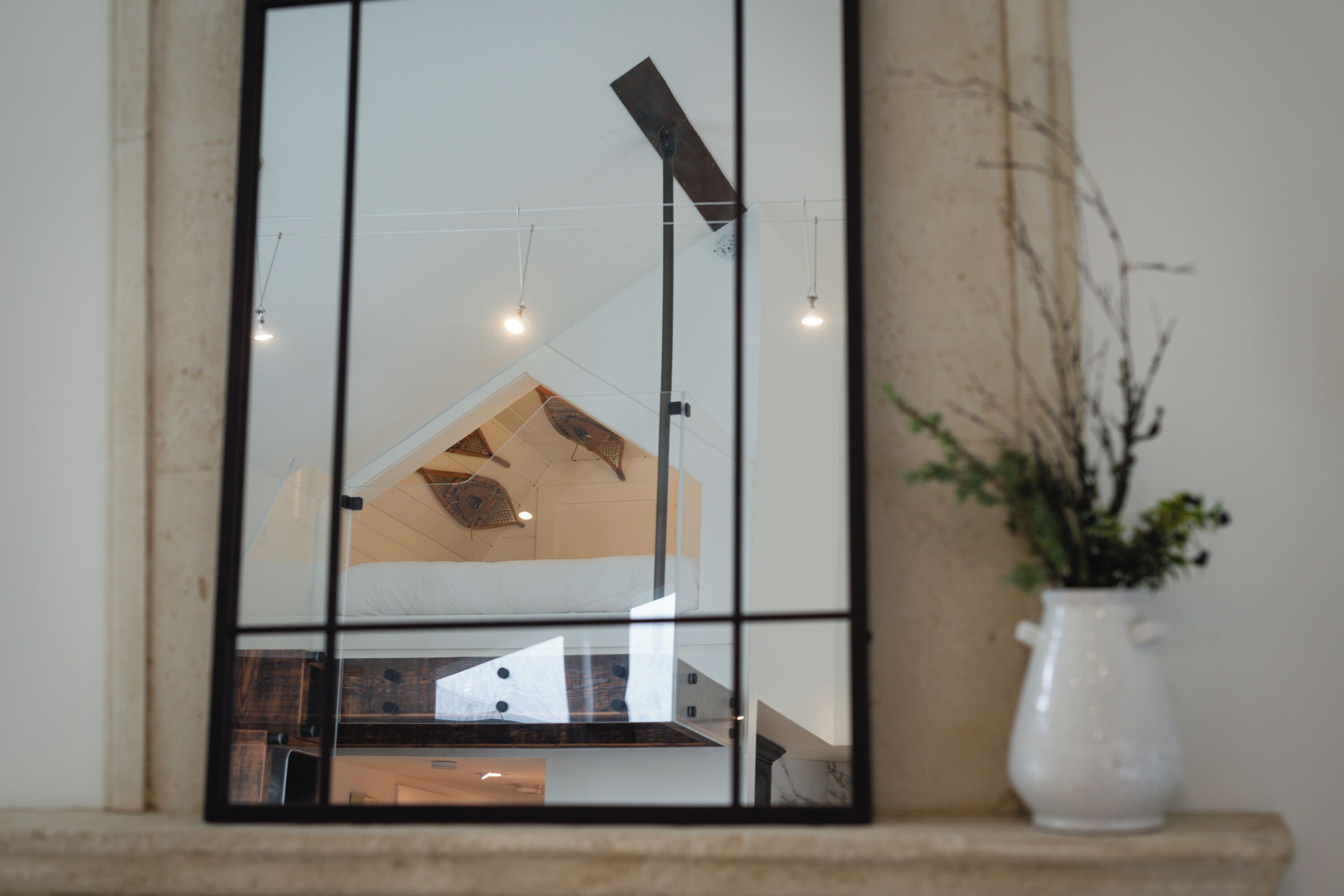 Un espejo refleja un interior moderno con una zona de cocina, una mesa de comedor y un jarrón blanco con follaje sobre un alféizar de piedra.