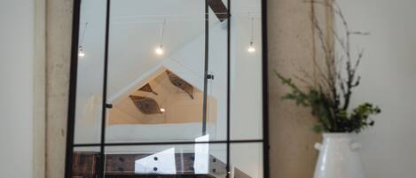 Un espejo refleja un interior moderno con una zona de cocina, una mesa de comedor y un jarrón blanco con follaje sobre un alféizar de piedra.