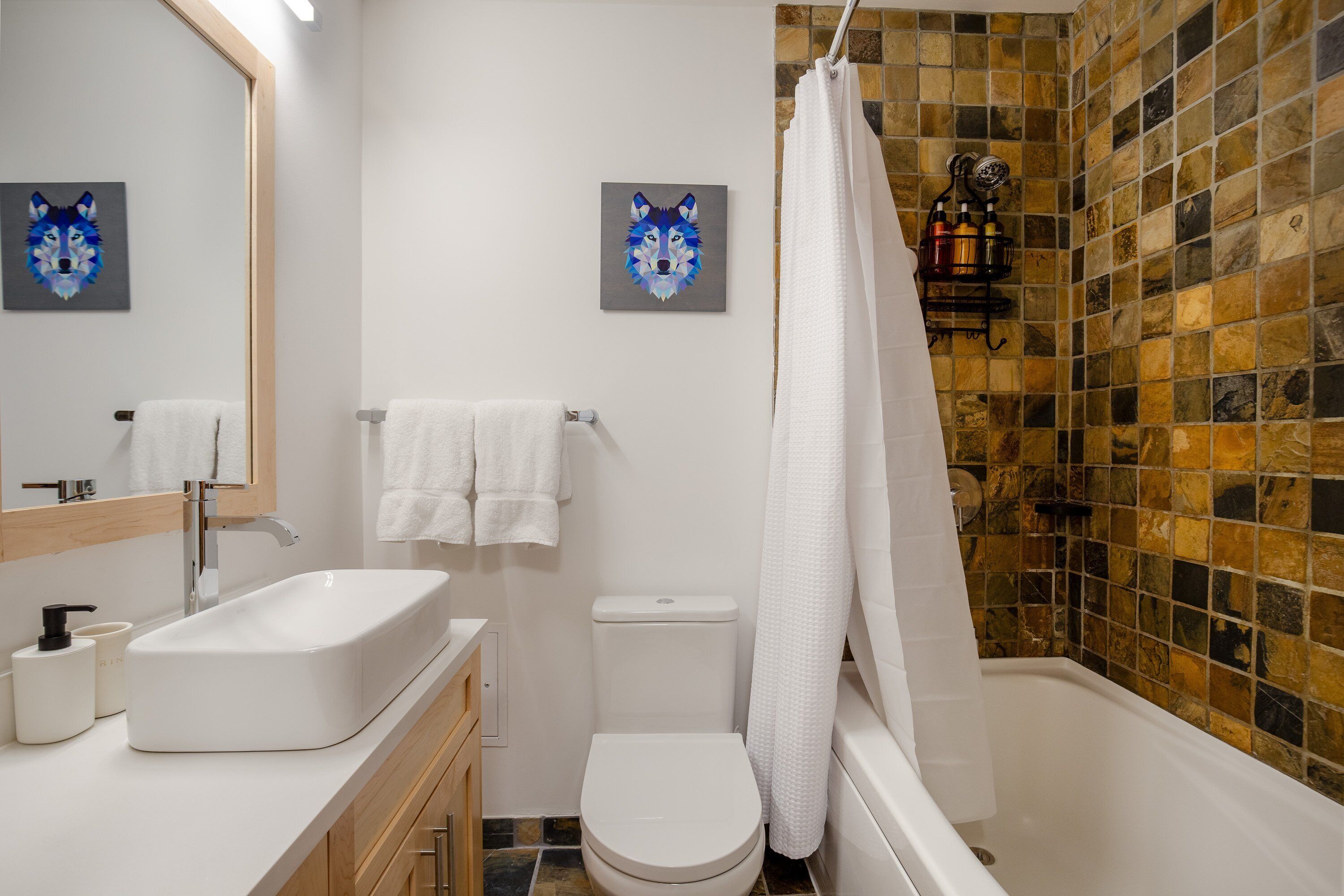A bathroom with a white sink, toilet, and bathtub, a shower curtain, and two wolf-themed paintings on the wall.