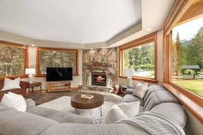 A living room with a stone fireplace, large windows, and a flat-screen TV.