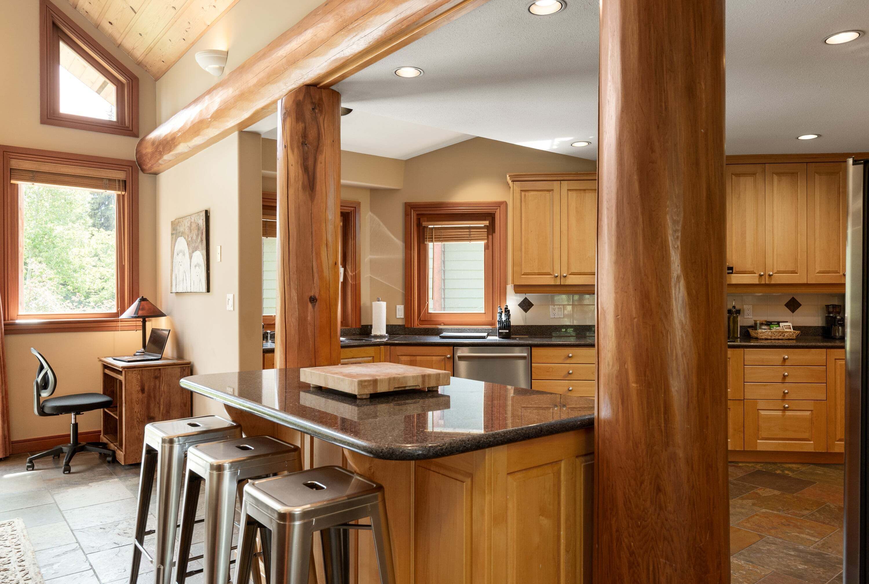A kitchen with wooden cabinets, a bar area with stools, a laptop on a small table, and a window with blinds.