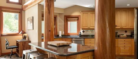 A kitchen with wooden cabinets, a bar area with stools, a laptop on a small table, and a window with blinds.