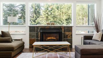 A living room with a fireplace, two sofas, a coffee table, and large windows with a forest view.