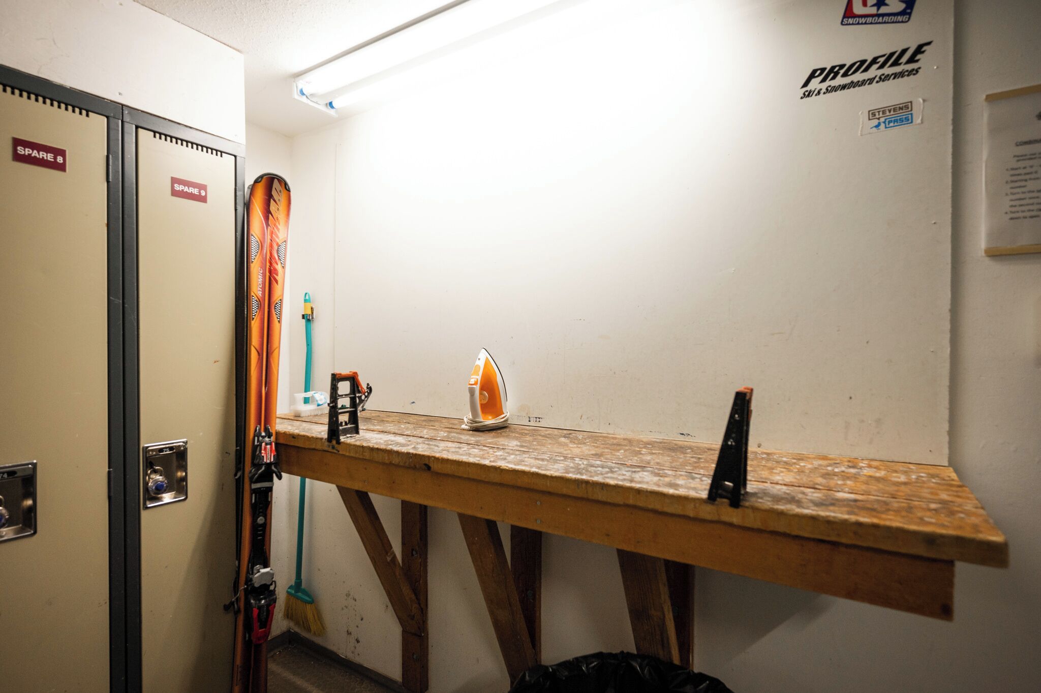 Une pièce avec un établi en bois, des paires de skis appuyés contre le mur et une enseigne indiquant « PROFILE, services d’entretien et de réparation de skis et de snowboards ».