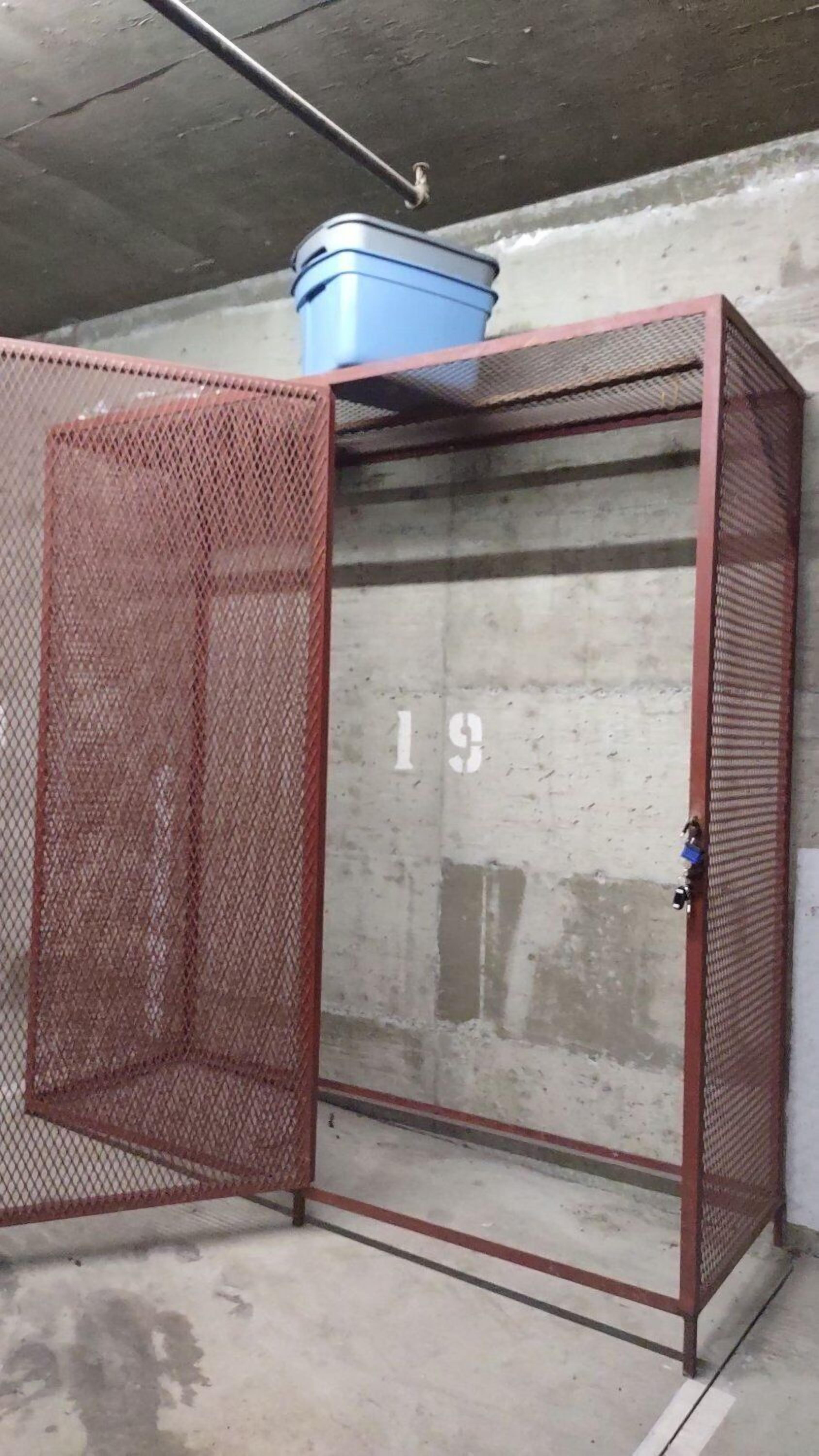 Un armario metálico con puerta de malla, un contenedor azul encima y el número '19' en la pared.