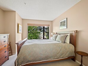 Een slaapkamer met een bed, een dressoir, een lamp en een raam met uitzicht op bomen.