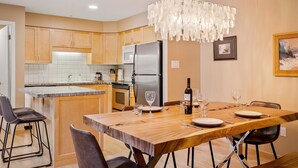 A modern kitchen with wooden cabinets, a stainless steel refrigerator, and a dining area with a wooden table and chairs. - Glacier Suite at Snowberry (Whistler)