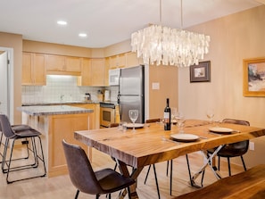 A modern kitchen with wooden cabinets, a stainless steel refrigerator, and a dining area with a wooden table and chairs. - Glacier Suite at Snowberry (Whistler)
