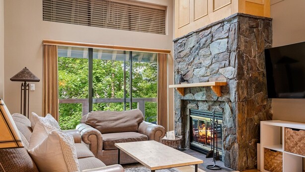 A modern living room with a stone fireplace, leather sofa, and a large window offering a view of greenery. - Glacier Suite at Snowberry (Whistler)