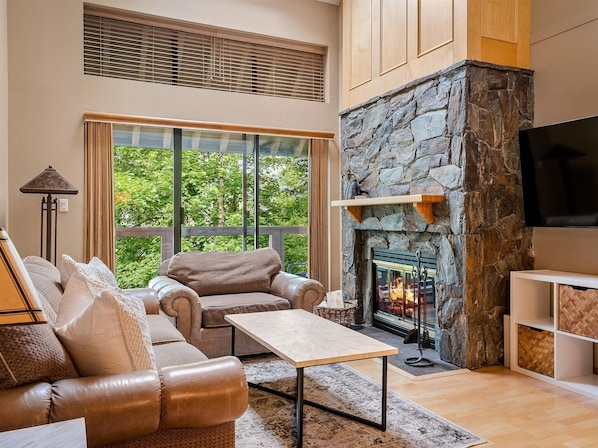 A living room with a stone fireplace, leather sofa, and a large window overlooking greenery. - Glacier Suite at Snowberry (Whistler)