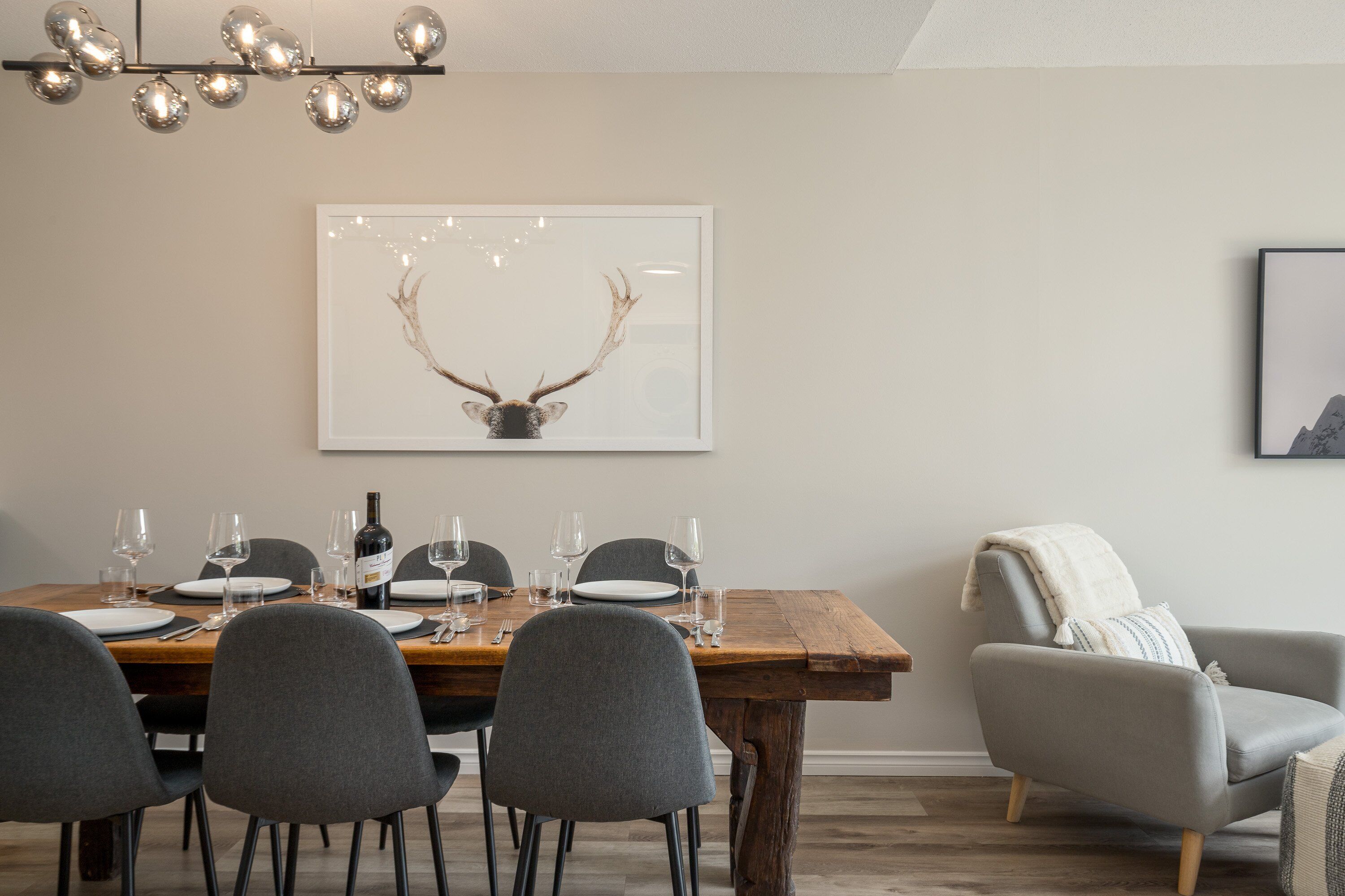 Un coin repas avec une table en bois dressée pour quatre personnes, des chaises grises, un lustre et des œuvres d’art encadrées accrochées au mur.