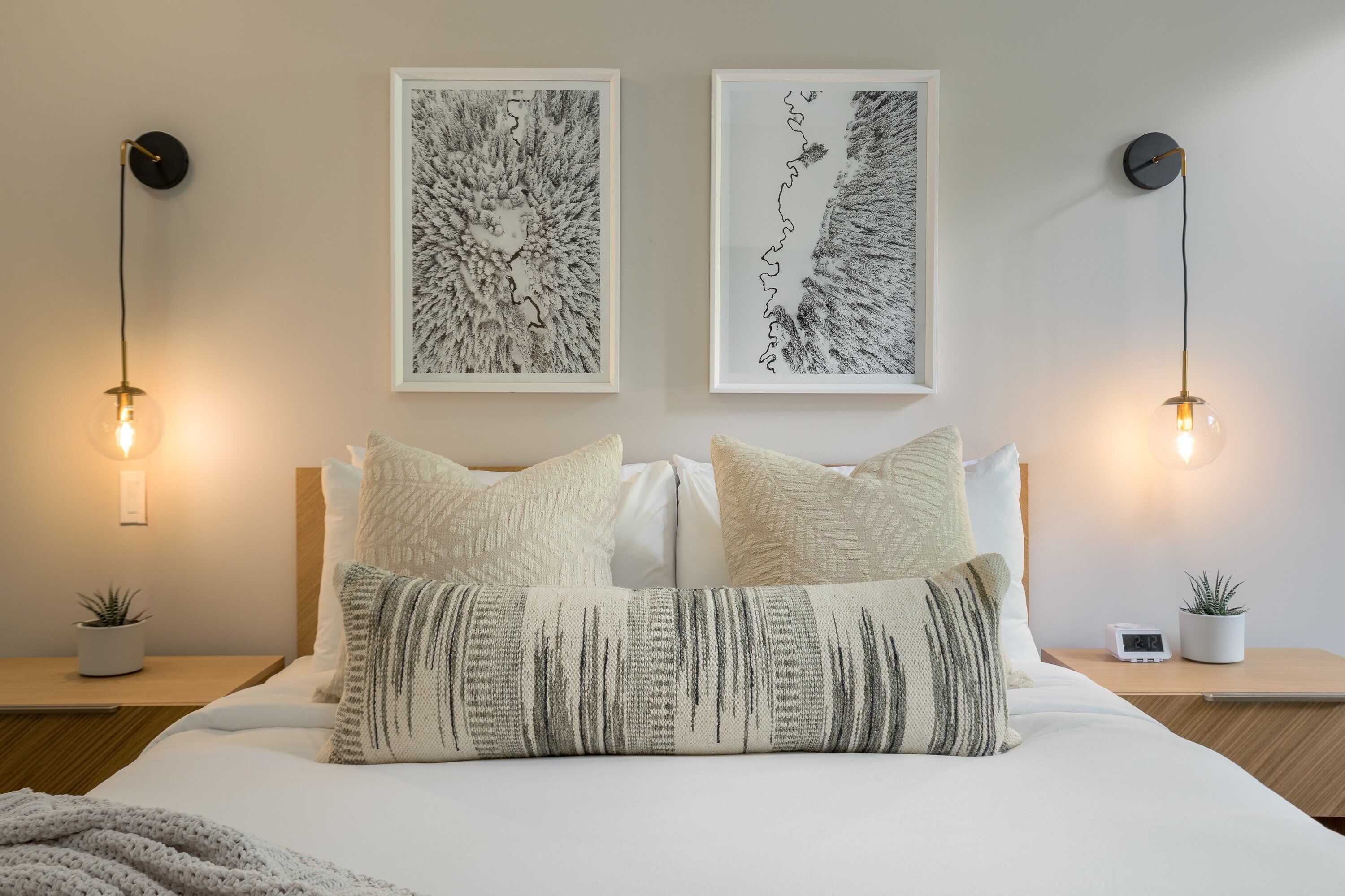 Een netjes ingerichte slaapkamer met een bed, twee wandlampen, twee ingelijste kunstwerken en een klein digitaal klokje op het nachtkastje.