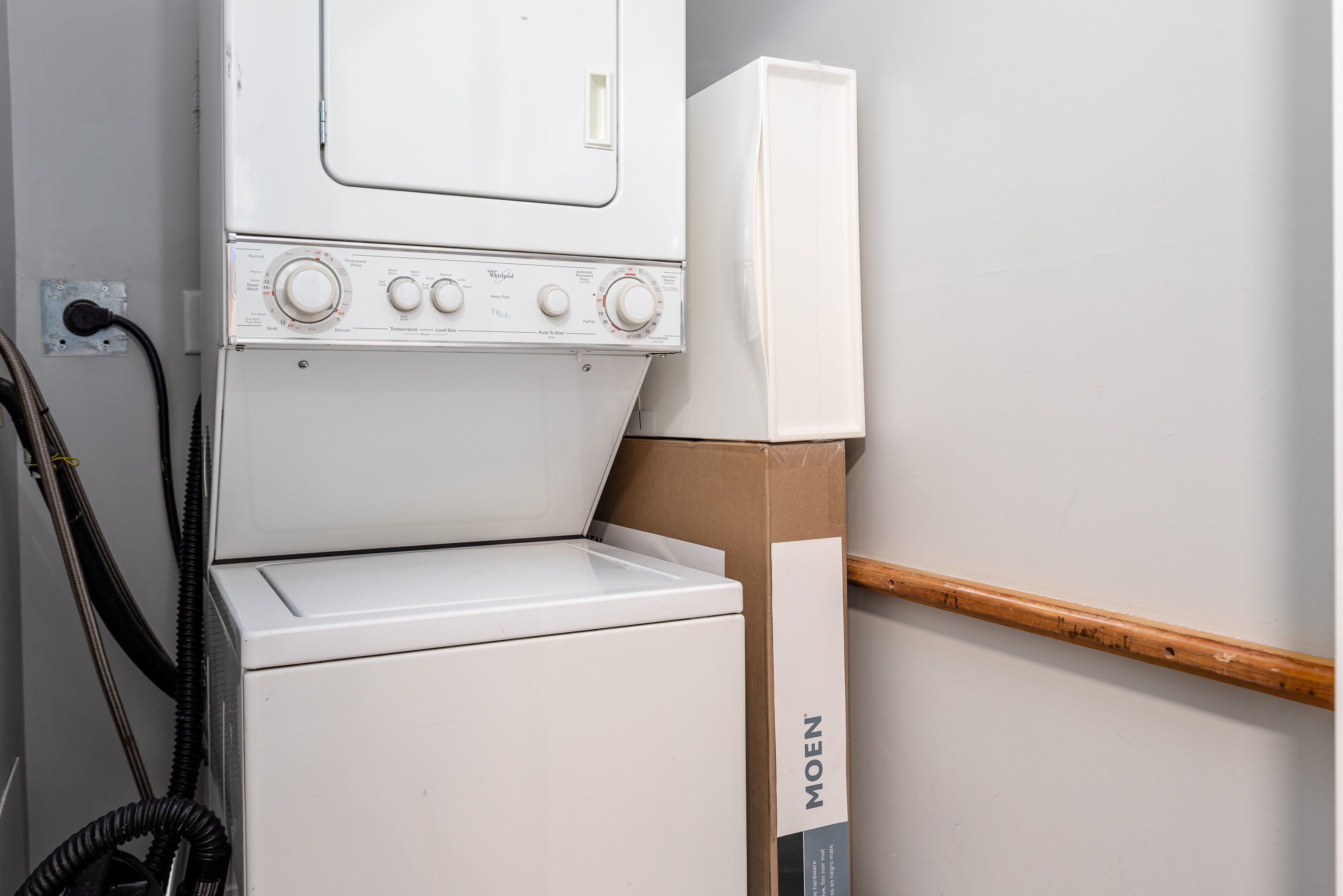 A white washing machine with control knobs and a hose, a wooden beam, and a cardboard box labeled 'MOEN' against a wall.