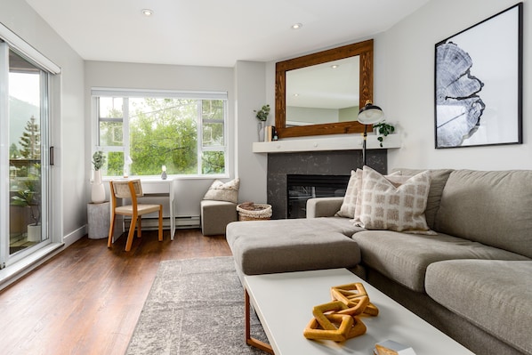 A modern living room with a sofa, a coffee table, a fireplace, and a dining area with a chair and table. - Misty Mountain at Marketplace Lodge (Whistler)