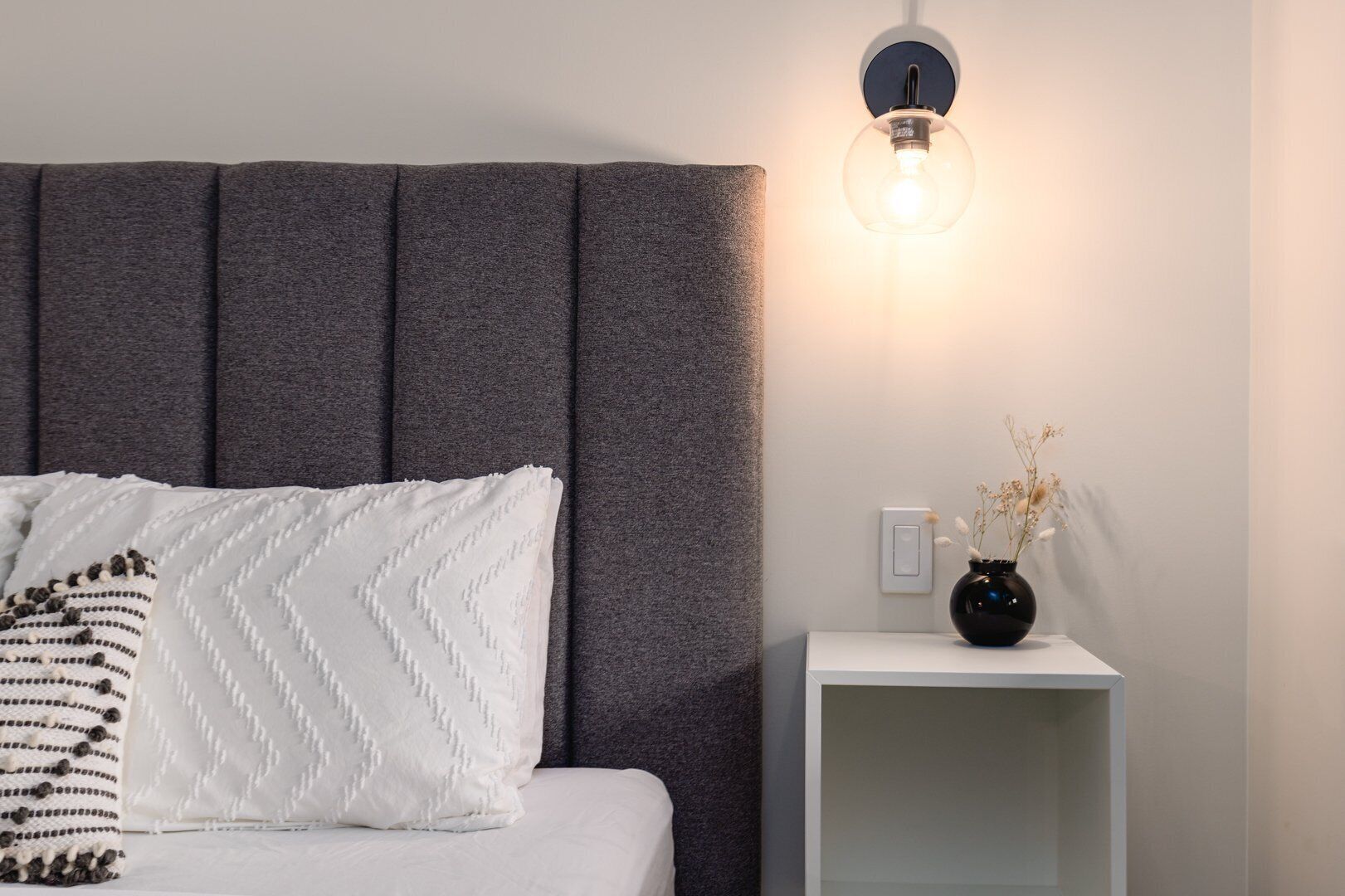A bed with a grey headboard, a white pillow with a zigzag pattern, a bedside table with a vase of dried flowers, and a wall-mounted light fixture.