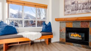 A cozy room with a bench, a fireplace, and a large window with a view of trees and mountains. - Mountain View Suite at Marketplace Lodge (Whistler)