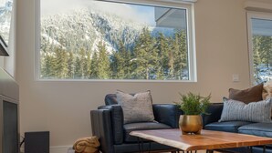 A modern living room with a large window, a black leather sofa, a wooden coffee table, and a fireplace. - Nature's Nook at Wedgewoods (Whistler)