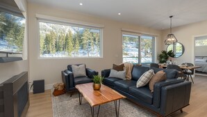 A modern living room with a sofa, coffee table, and large windows offering a view of a snowy landscape. - Nature's Nook at Wedgewoods (Whistler)
