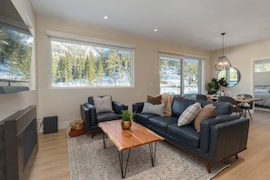 A modern living room with a sofa, coffee table, and large windows offering a view of a snowy landscape. - Nature's Nook at Wedgewoods (Whistler)
