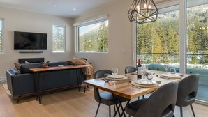 A modern living room with a sofa, TV, dining table set for four, and large windows offering a mountain view. - Nature's Nook at Wedgewoods (Whistler)