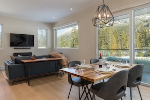 A modern living room with a sofa, TV, dining table set for four, and large windows offering a mountain view. - Nature's Nook at Wedgewoods (Whistler)