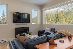 A modern living room with a fireplace, a flat-screen TV, and large windows offering a forest view. - Nature's Nook at Wedgewoods (Whistler)